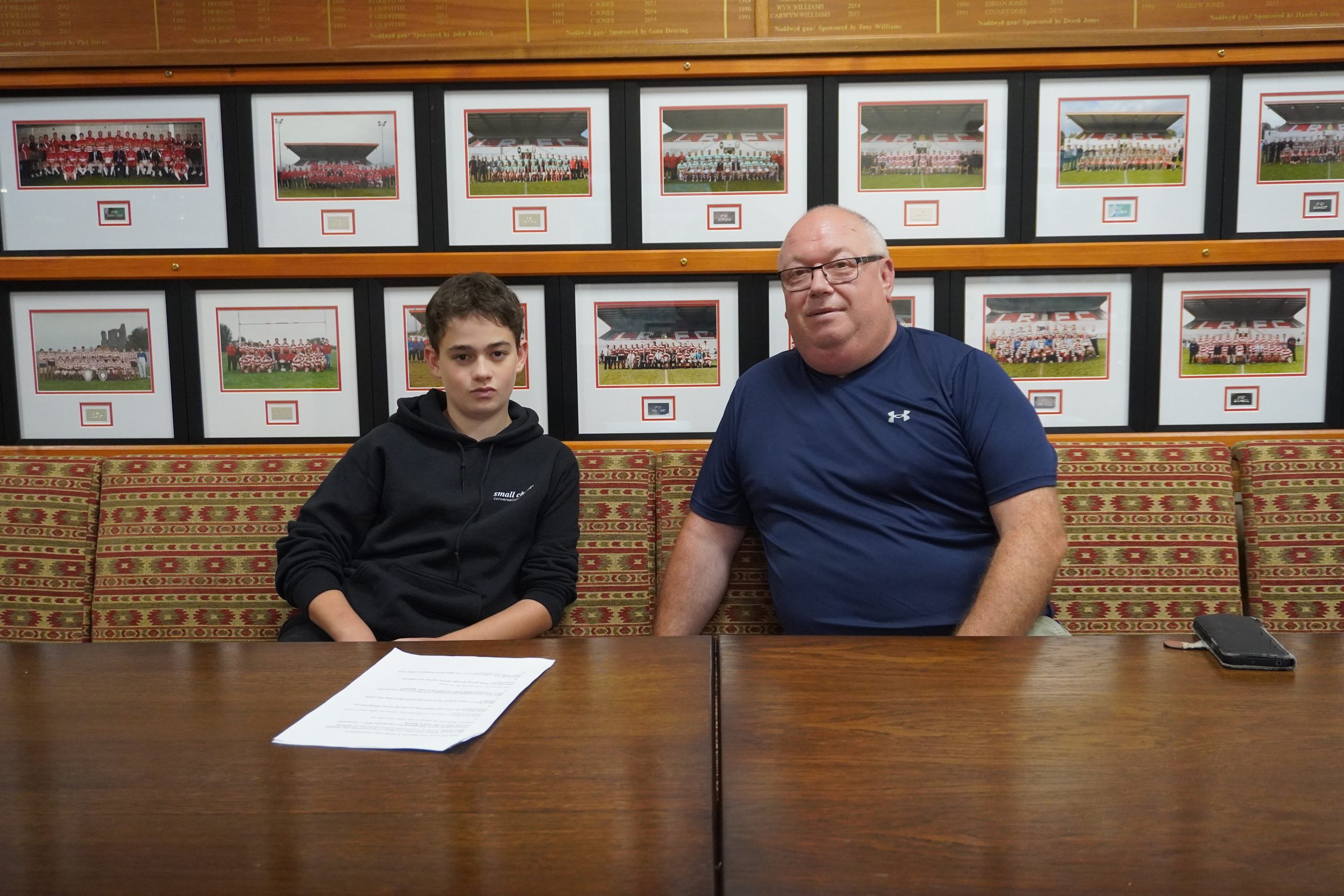 András seated beside Andrew “Cantona” Bryn-Jones at Llandovery RFC, with historic team photographs displayed behind them.
